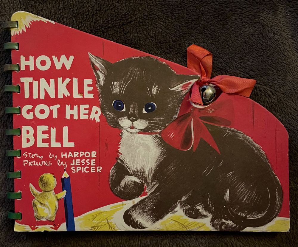 Photo of a book cover with an illustration of a shorthaired black tuxedo kitten with a red ribbon around its neck, which overlaps with a real bell and ribbon the are attached to the book. There is also a little chick with a pencil and the words “HOW TINKLE GOT HER BELL
Story by HARPOR
Pictures by JESSE SPICER”