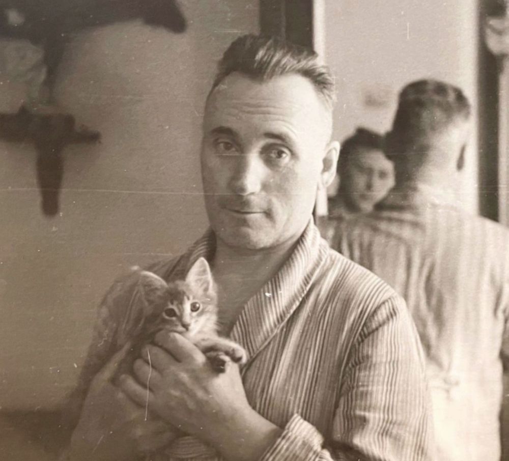 Black and white photo of a handsome white man wearing pajamas and smiling a little as he holds up a tabby kitten. Both man and kitten are looking at the camera. Another person can be seen in the mirror behind the man.