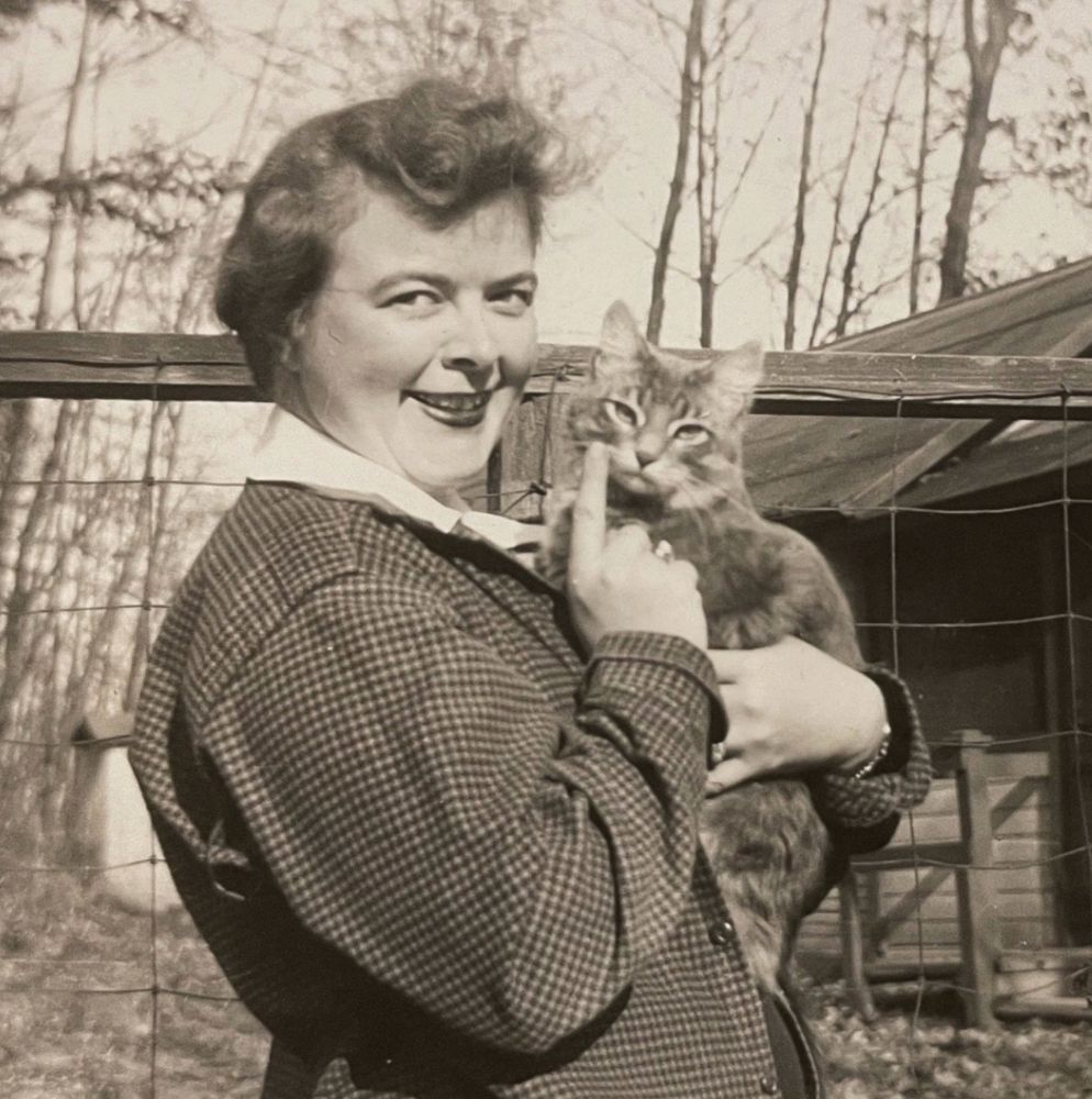 Black and white photo of a white woman in a dark checkered coat, smiling at the camera and pointing to the beautiful medium haired tabby cat she cradles in her arms. The cat also looks at the camera.