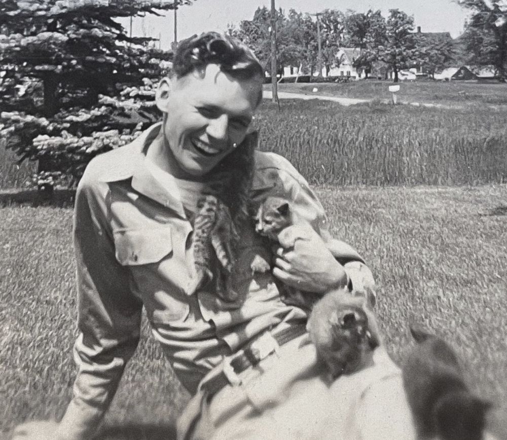 Black and white photo of a young white man sitting on a lawn in bright sunlight, smiling broadly at the camera. He has a tortie kitten in his left hand white a tabby climbs up his shirt next to his neck, and two more kittens are wobbling around on his outstretched legs.
