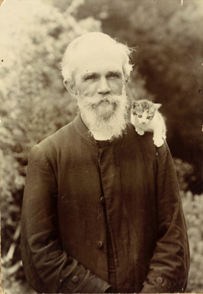 Black and white photo of a smiling, very kind looking older man with white beard, standing outside and wearing black priest's garments. On his left shoulder is perched a tiny shorthaired white and tabby kitten.