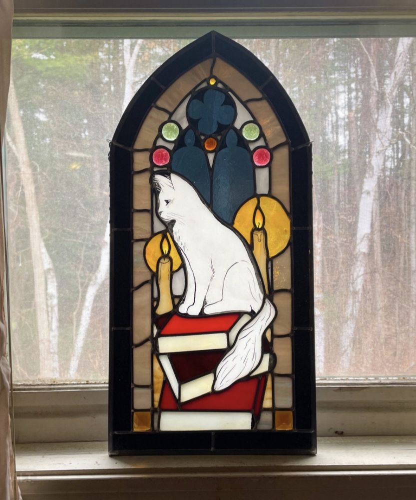 A stained glass piece about 2 ft tall and 1 ft wide, depicting a medium haired white cat sitting on top of a stack of large hardcover books, two off white candles glowing nearby. In the background you see tall, arched church windows with a round window above them. The glass set in the windows is a mellow dark blue like the night sky. That in combination with the candles gives the scene a feeling of cozy nighttime.