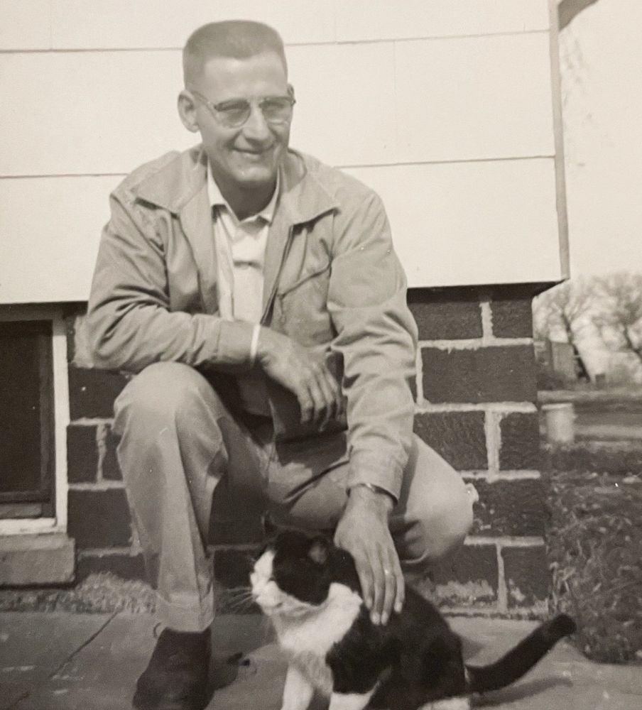 Black and white photo of a white man with a crew cut kneeling on a sidewalk and smiling at the camera while petting a happy looking shorthaired black tuxedo cat.