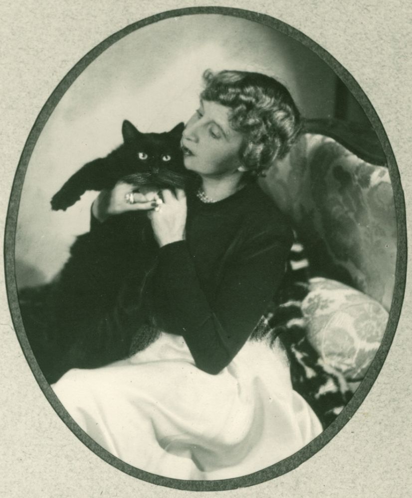 Black and white photo of a white woman with curly blonde or light brown hair, sitting on an upholstered, fancy looking chair and holding a large cat up by its armpits to affectionately snuggle it. The cat looks displeased but tolerant of this.