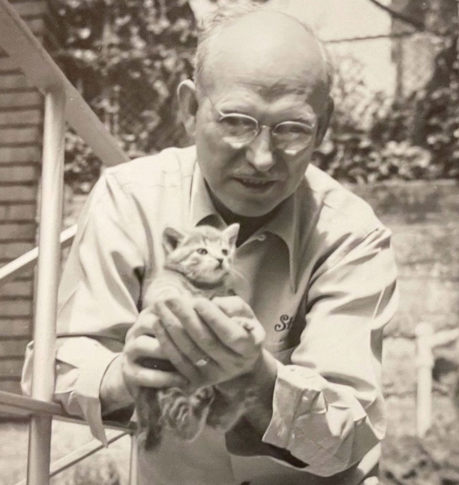 Black and white photo of a kind looking older white man wearing glasses, gently holding a very brave looking tabby kitten up to the camera.