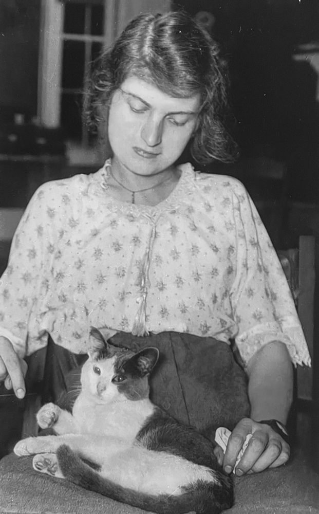 Black-and-white photo of a light skinned young woman sitting up in a chair. She is looking down at a short haired, white and gray cat. She’s also holding out one finger as if playfully teasing the cat with it.