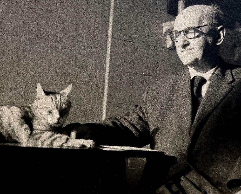 Black and white photo of a balding elderly white man in a suit, sitting at a table and looking affectionately at a tabby cat lying next to him. He also seems to have one hand stretched out to touch the cats’ paw.