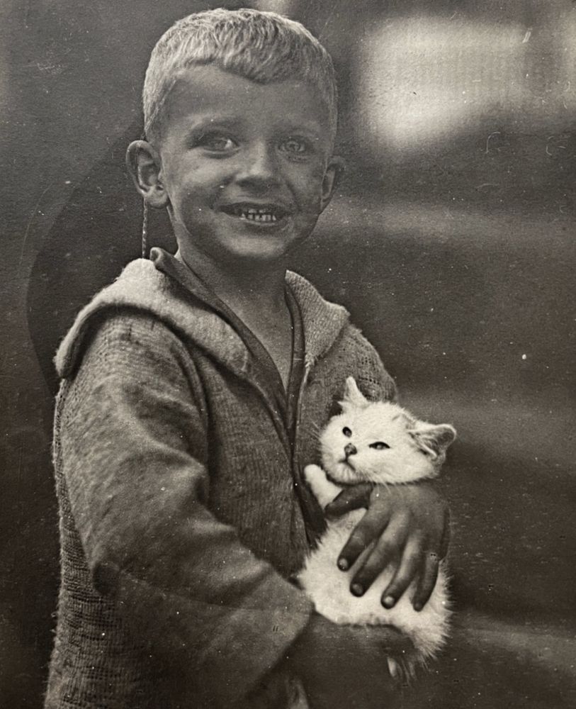 Black and white photo of a young white boy with blonde hair, posing for the camera with a smile. In his arms is a poofy white kitten which doesn’t look like it is enjoying this at all.