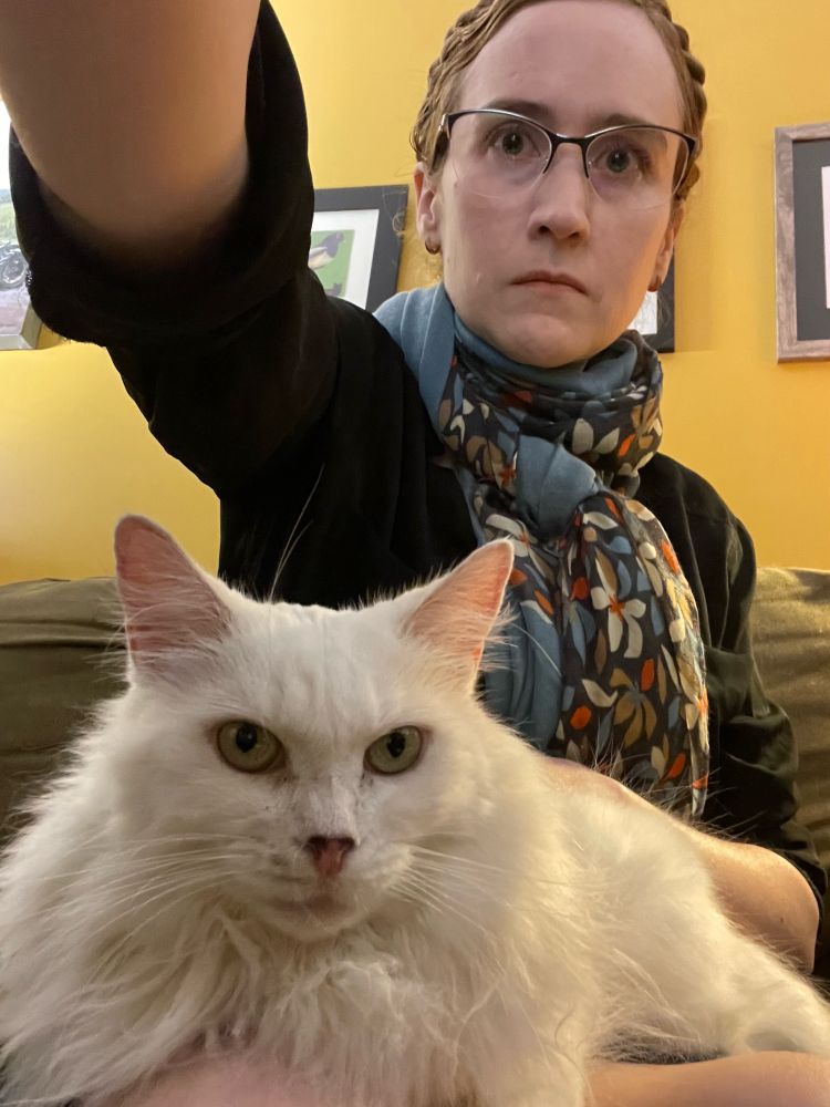 Photo of a 46 year old white woman with glasses and a braid crown and a longhaired white cat in her lap.