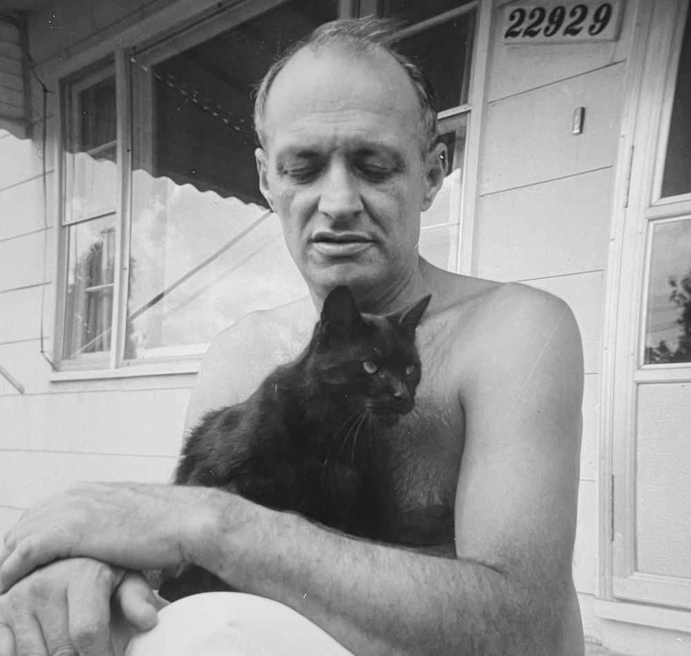Black and white photo of a balding, white, shirtless man sitting on the stoop of a white house. He is looking affectionately down at a shorthaired black cat sitting contentedly in his lap.