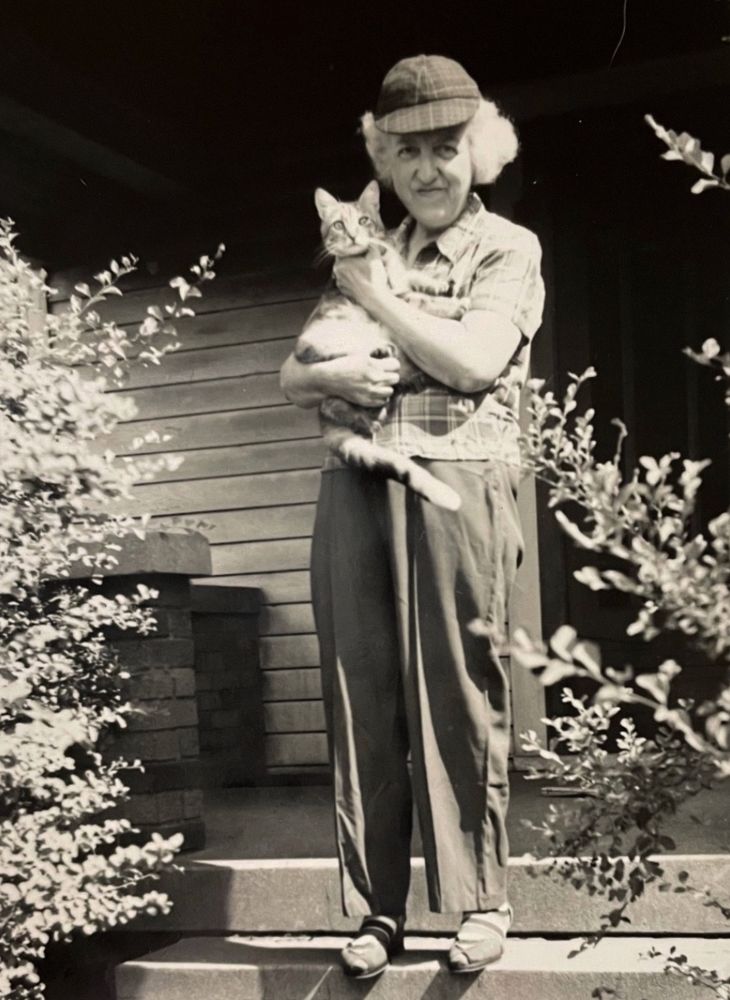 Black and white photo of an older white lady standing on a house porch wearing pants, a plaid blouse, and her white hair puffing out from under a plaid cap. She is smiling at the camera and cradling a shorthaired tabby cat in her arms.