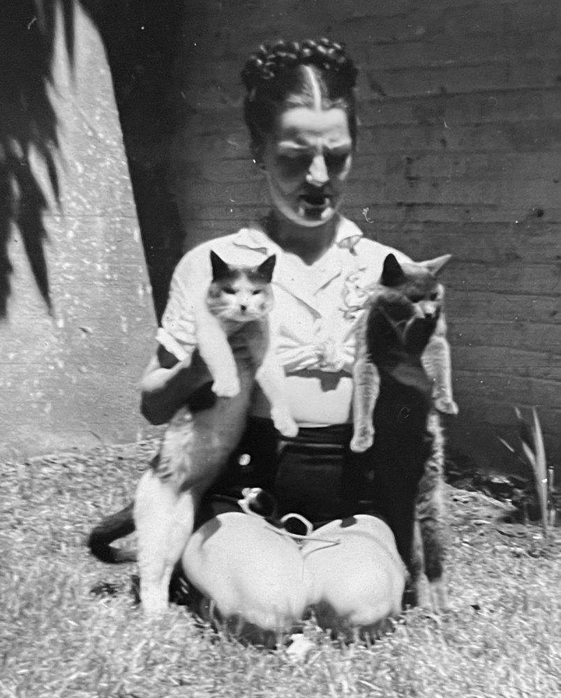 Black and white photo of a light skinned woman kneeling on a lawn in the sun. She’s holding up two shorthaired cats, one under each arm, as if showing them off.