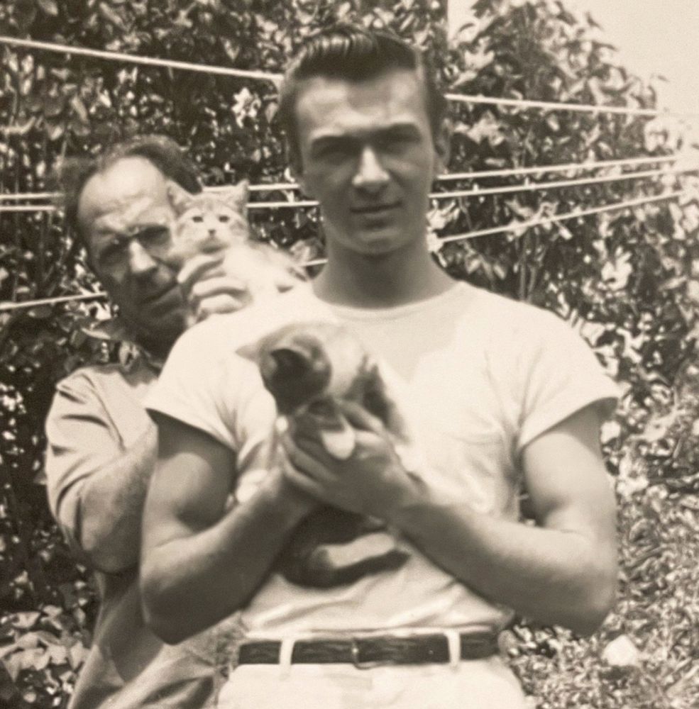 Black and white photo of a muscular young white man with dark, slicked back hair, staring at the camera and holding up a shorthaired tuxedo tabby kitten. An older white man behind him props another kitten up on his shoulder.