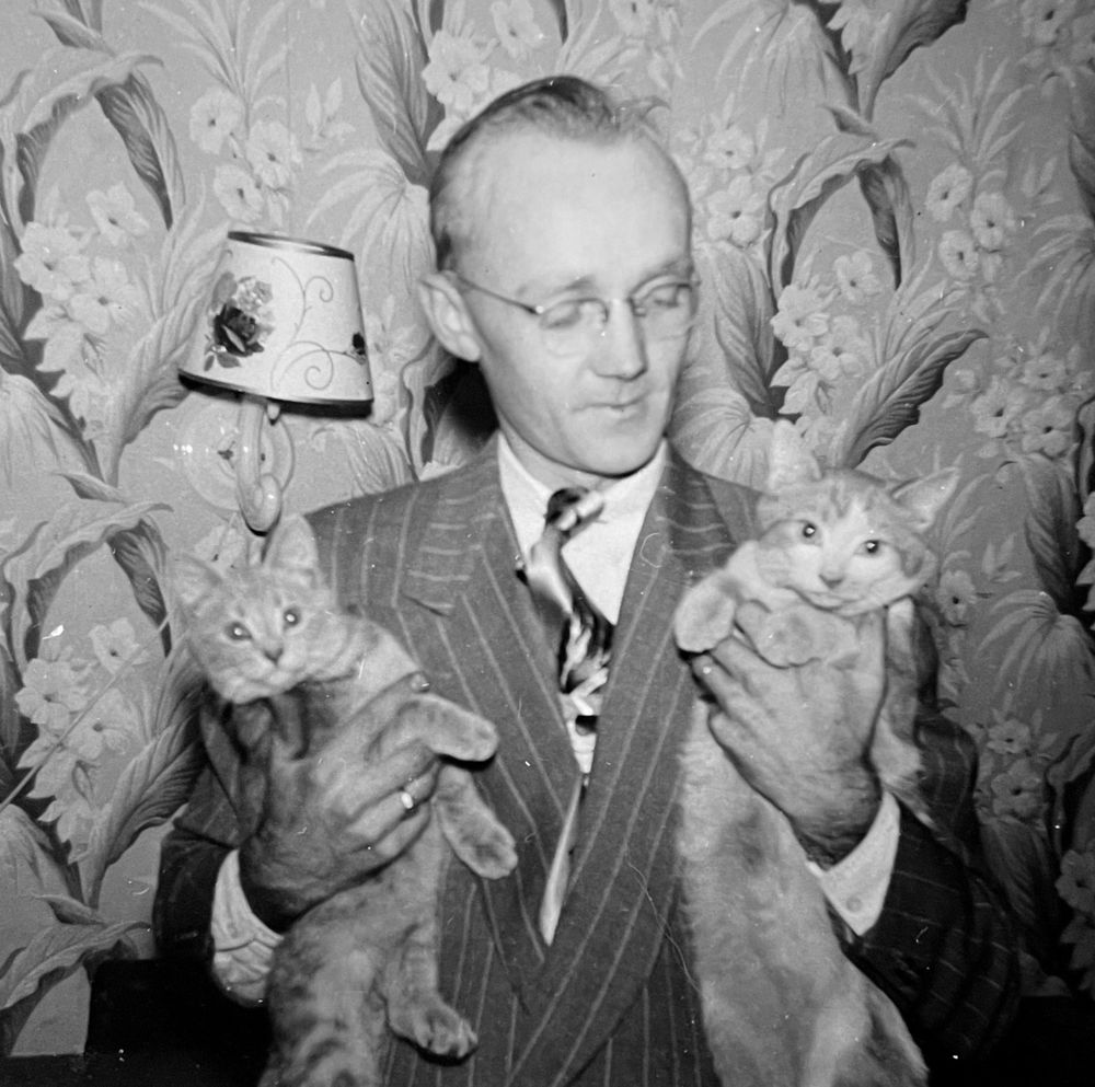Black and white photo of a white man in wide lapel pinstriped suit, looking affectionately down at two shorthaired tabby cats that he holds under his arms. He’s in a living room with bold floral wallpaper.