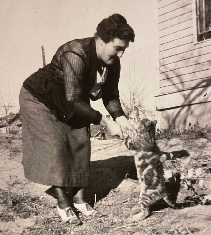 Black and white photo of a stout middle aged woman with dark hair standing outside on a scrubby dead lawn. She is leaning forward and holding the forepaws of a shorthaired tabby cat so that it is “dancing” with her on its hind legs. She seems to be smiling at the cat and maybe talking to it.