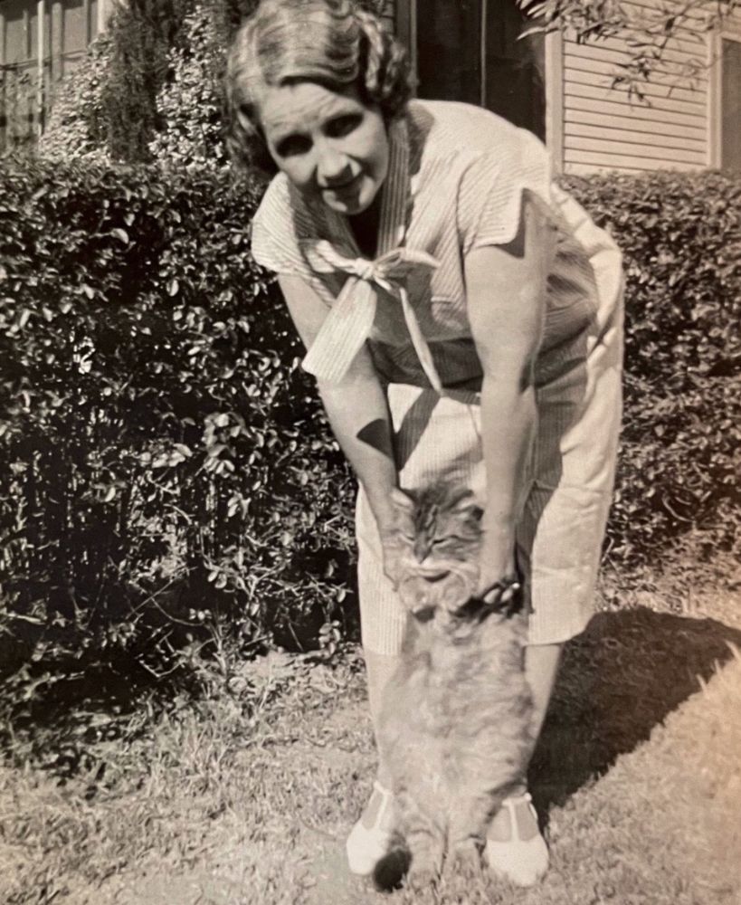 Black and white photo of a white woman standing outside on a lawn in front of a hedge, holding a fluffy tabby cat up by its elbows so that it is standing on its hind legs. She looks like she’s being quite gentle but the cat looks disgusted regardless.