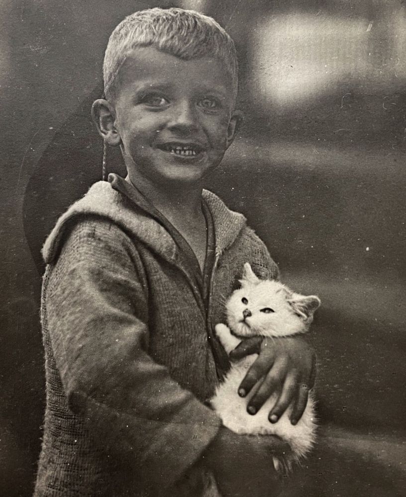 Black and white photo of a young white boy with blonde hair, posing for the camera with a smile. In his arms is a poofy white kitten which doesn’t look like it is enjoying this at all.