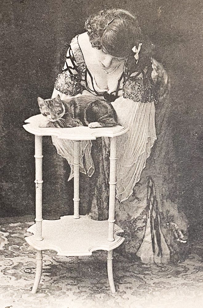 Black and white photo of a light skinned woman skinned woman in an elaborate beaded and layered gown, leaning forward and adoringly snuggling a shorthaired tabby on a small white table.