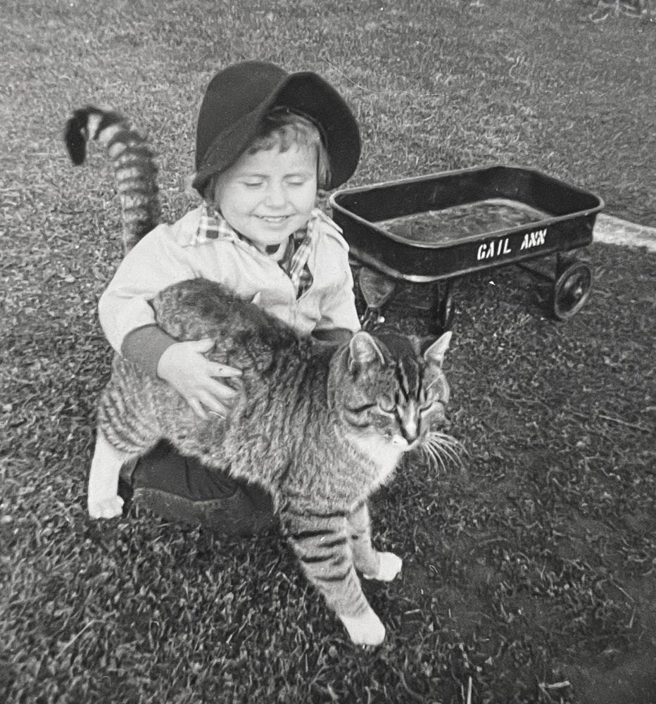 Black and white photo of a little white girl kneeling on grass outdoors next to a toy wagon with “Gail Ann” stencil painted on it. She is smiling and hugging a big shorthaired tuxedo tabby cat who looks very happy standing beside her.