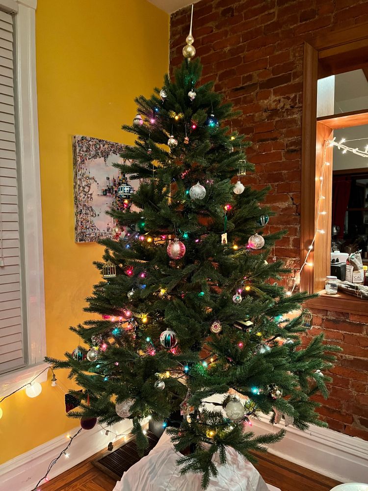 Classic Christmas tree with a white skirt in a corner against a yellow wall and a brick kitchen pass-through. A near-perfect balance of antique glass balls, hanging doodads, and stuffed llama.