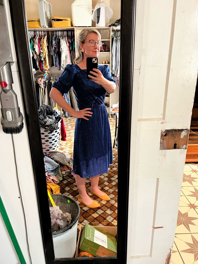 A white woman takes a full-length selfie, wearing a very 1983 blue polyester dress, round earrings, and hair pinned back. She’s channeling Martha from the Americans but apparently no one west of the Wabash watched that show. 