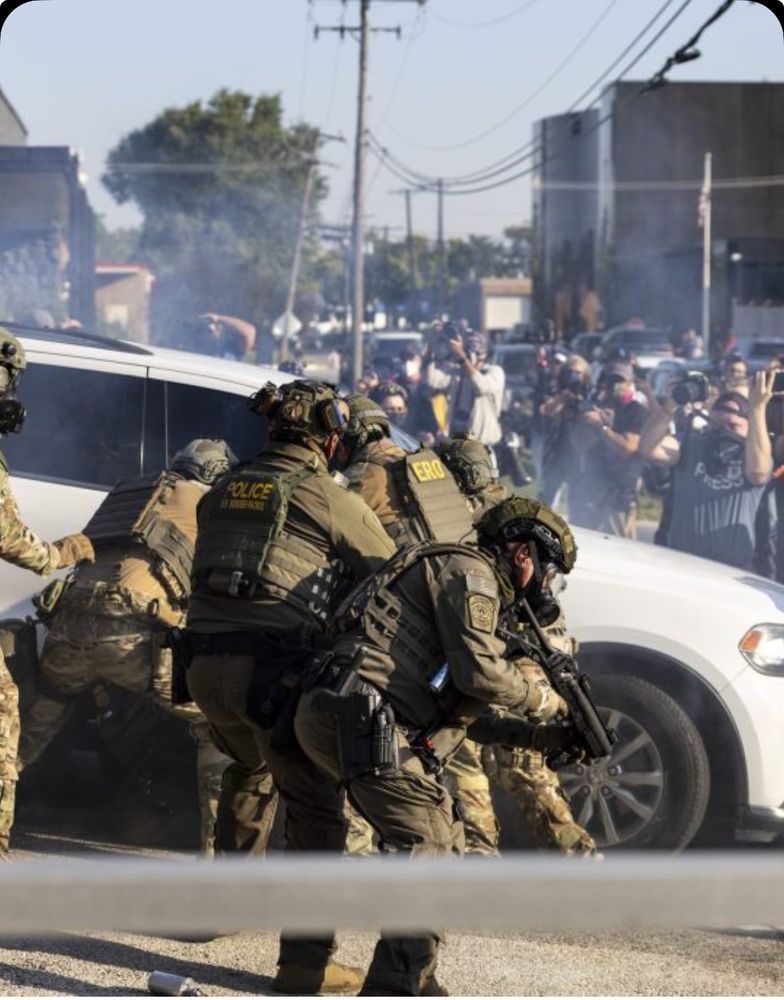 The image shows a tense law enforcement operation involving heavily armed officers in tactical gear, some marked with “POLICE U.S. IMMIGRATION” and “ERO” (Enforcement and Removal Operations). They appear to be surrounding a white SUV, with weapons drawn and smoke in the air. In the background, a crowd of onlookers and press with cameras are capturing the scene. The atmosphere suggests a high-stakes immigration raid or enforcement action, underscored by a visible media presence.