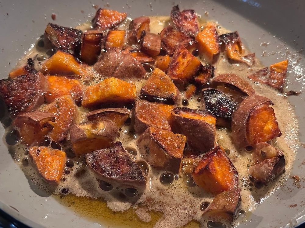 Close-up of cubed sweet potatoes with skins on, sautéing in a pan. The pieces are browned and slightly charred in spots, sitting in a bubbling buttery, caramel-like sauce that looks like it may include sugar or maple. Warm orange and deep brown tones suggest candied or pan-roasted sweet potatoes.