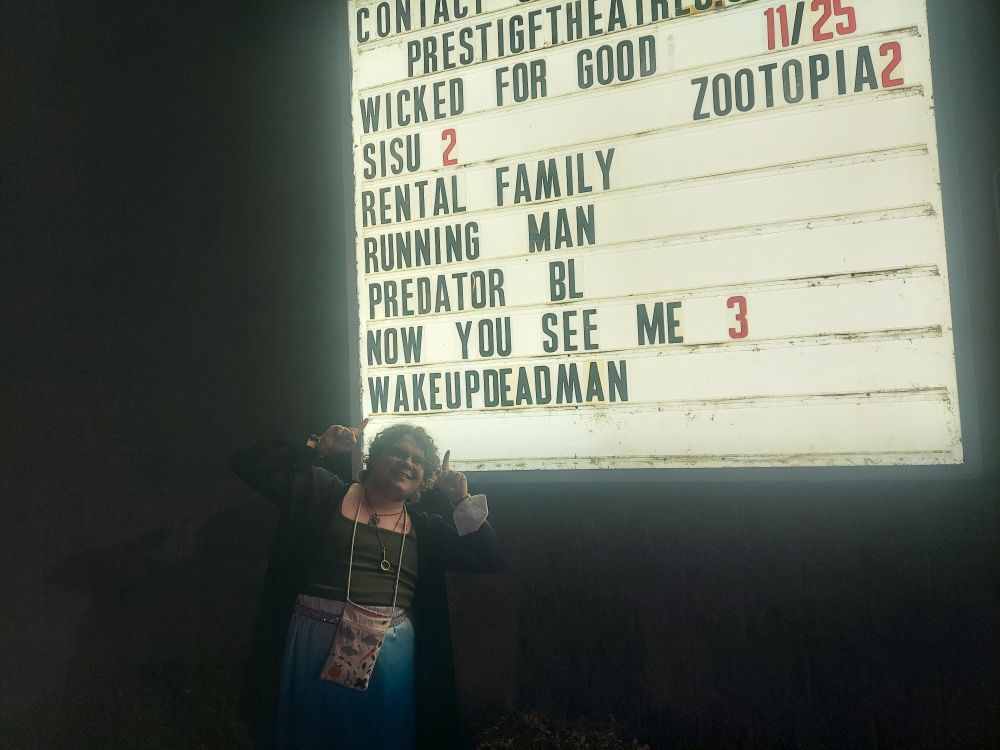 A photo of Casper smiling and pointing at a movie theater sign that reads a list of films, including "Wake Up Dead Man".