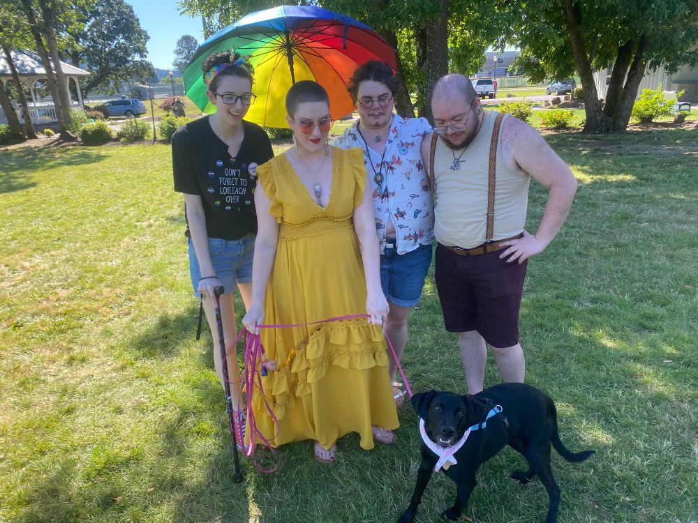 Hadrian and Kelsey from Fae Realm Productions standing next to Casper and Jason from Thesperience, all standing in front of a black dog on a bright pink leash. Casper is holding a rainbow umbrella to shield his friends from the sun.

The people are all looking at the dog with smiles or curious expressions as the dog looks up at the camera.