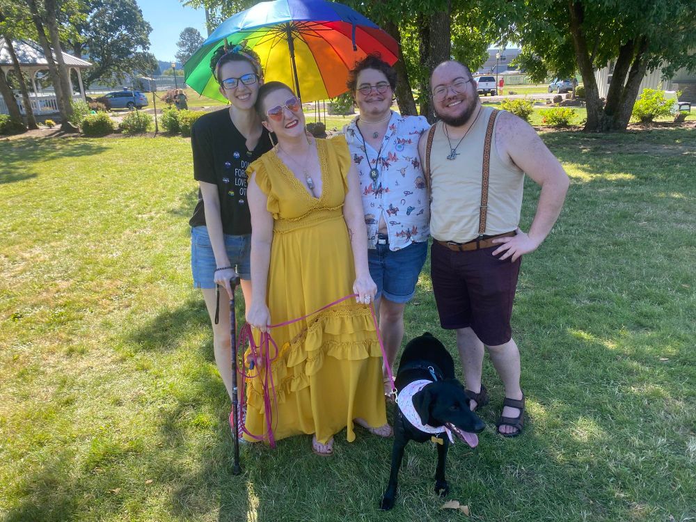 Hadrian and Kelsey from Fae Realm Productions standing next to Casper and Jason from Thesperience, all smiling and standing in front of a black dog on a bright pink leash. Casper is holding a rainbow umbrella to shield his friends from the sun.

The people are all looking at the camera as the dog looks at something off camera.