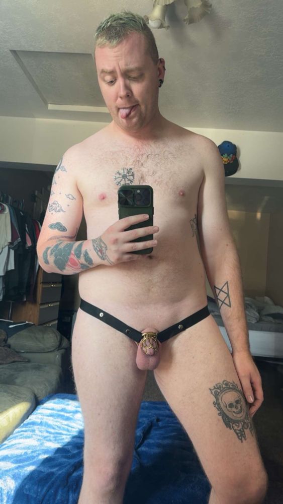 Banshee naked save for his gold chastity cage and black elastic strap for the cage, his tongue is sticking out 