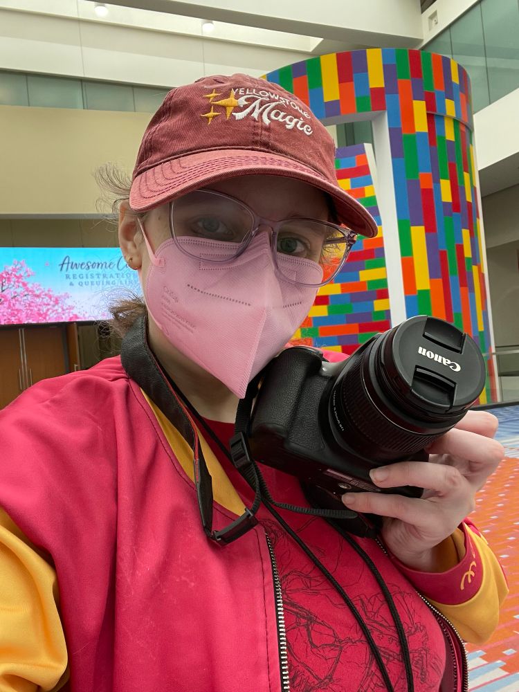 A selfie of Aviva, wearing a Yellowstone Magic baseball cap and bomber jacket, big light purple glasses, and a light pink mask She is holding a dslr camera. 