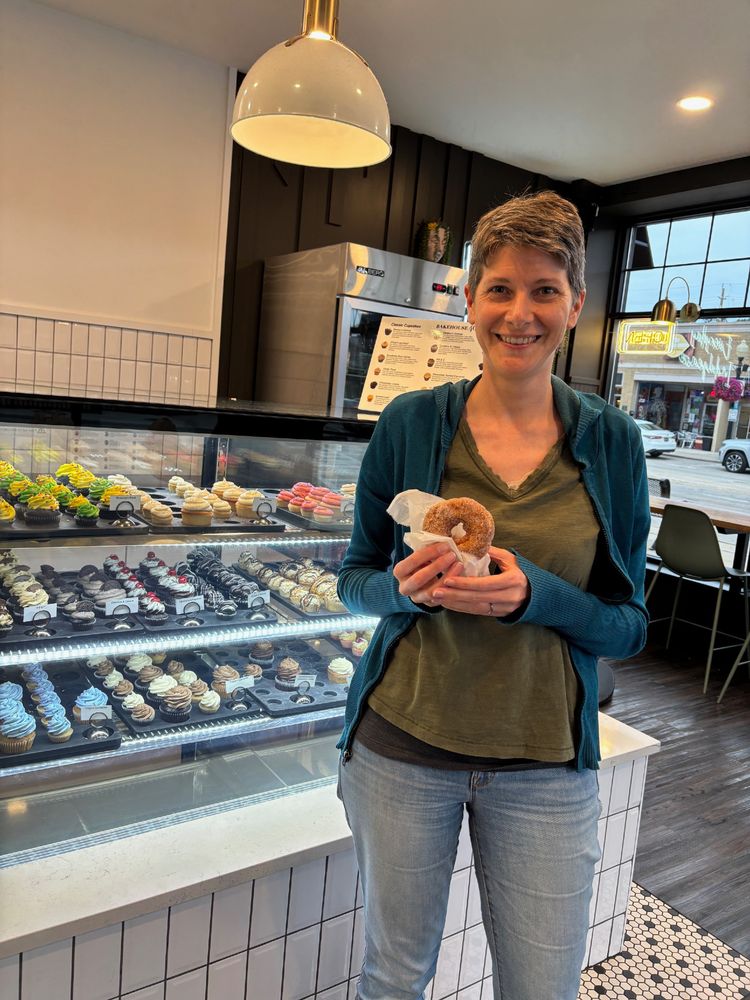 Quinn smiling and holding a donut. She is standing in front of a display case filled with cupcakes.