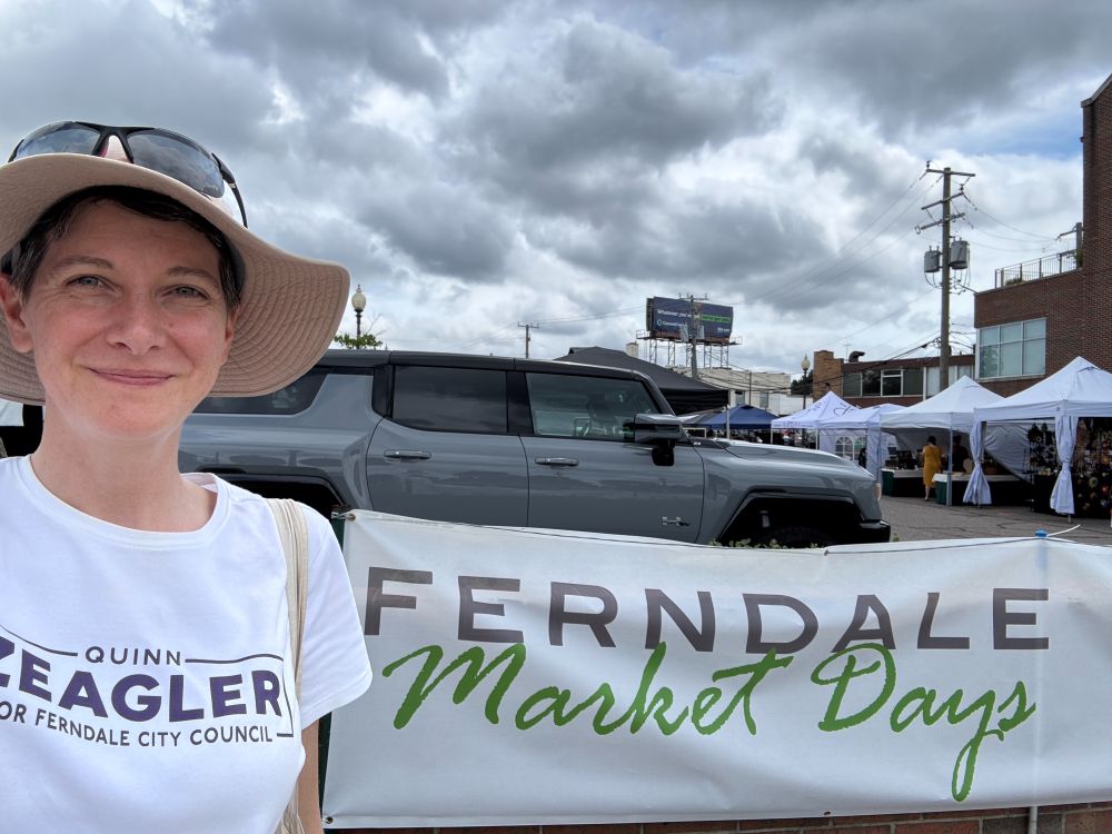 Quinn is smiling in front of a banner that reads "Ferndale Market Days." Vendor tents are in the background behind the banner.