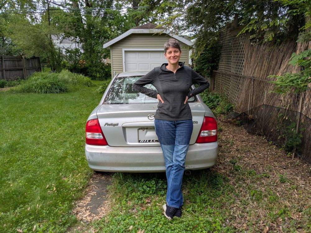 Quinn smiling in front of the closed trunk of a silver car. There is no license plate on the car.