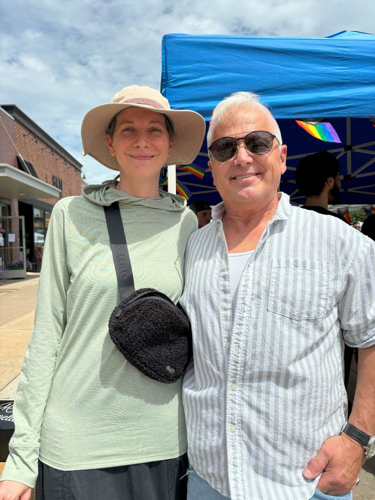 Two people smiling and standing in front of a booth with a Pride flag. Quinn is wearing a green shirt and sunhat. Dale is wearing a white shirt and sunglasses.
