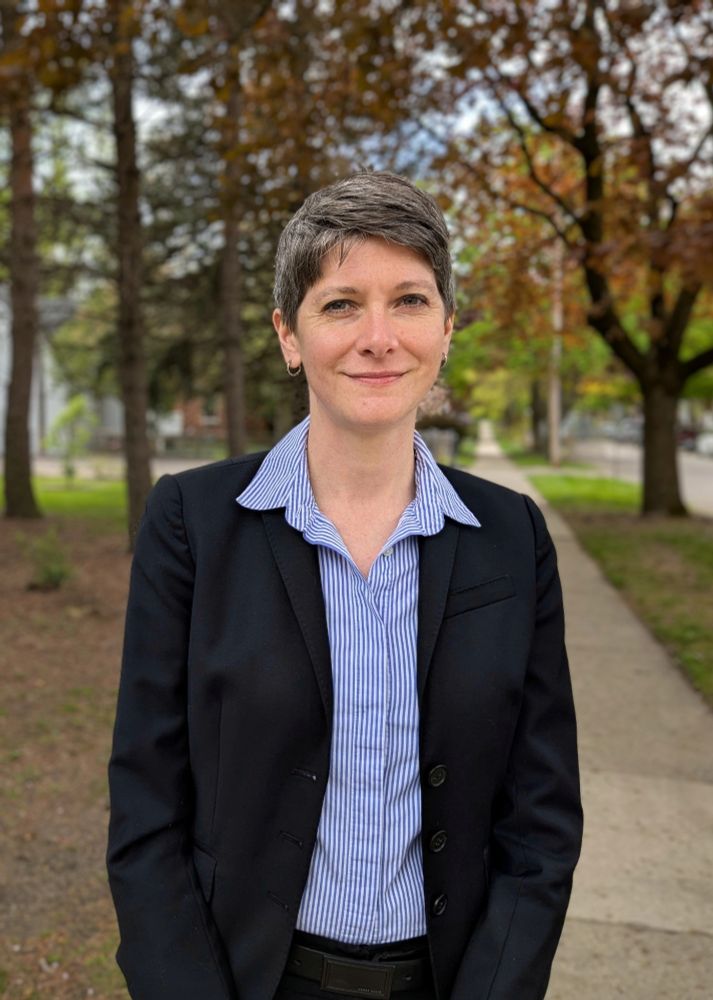 Quinn is smiling at the camera wearing a black work jacket and blue shirt.  Behind her are a sidewalk and trees.