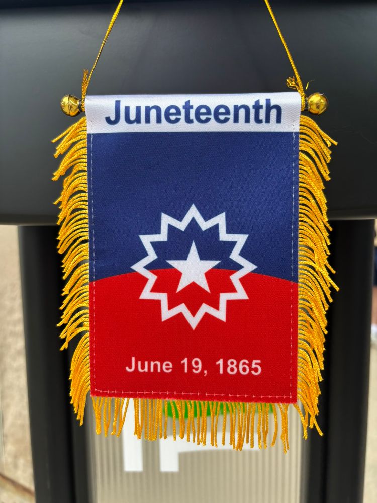 A small Juneteenth flag with yellow fringe. The flag reads Juneteenth June 19, 1865. The top of the flag is blue and the bottom of the flag is red. In the center is a white five point star surrounded by a white 12-point star.