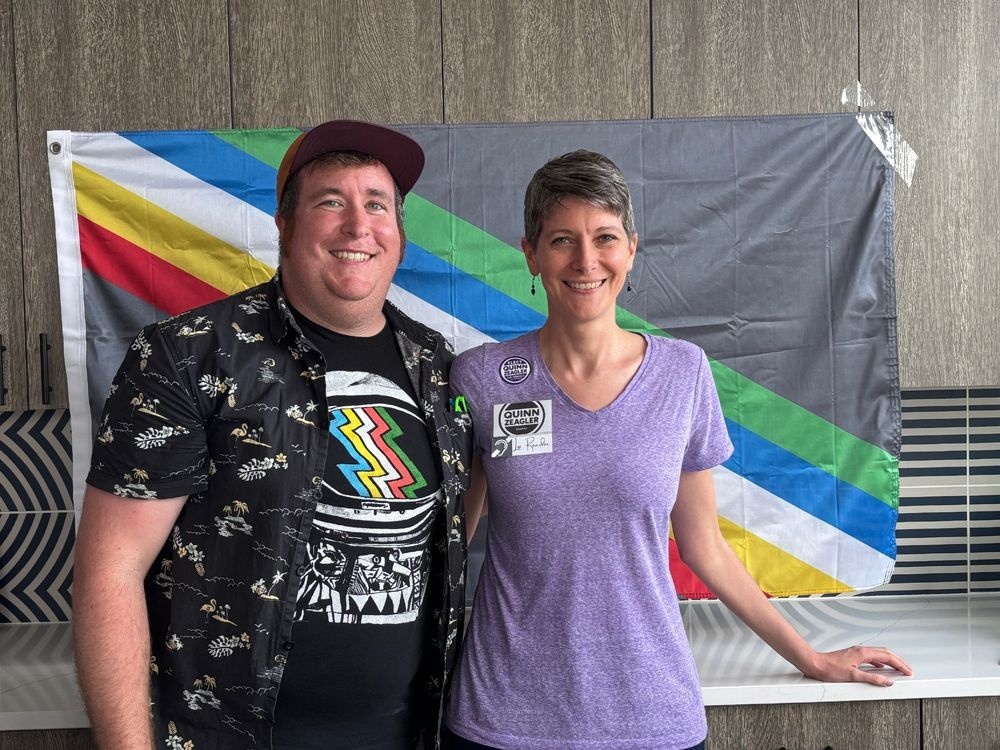 Alan Hejl and Quinn Zeagler are smiling in front of the Disability Pride Flag. The flag is five stripes going from the upper left to bottom right (from top to bottom, the stripes are green, blue, white, yellow, and red) on a gray background. Quinn's nametag identifies her as a lip reader.