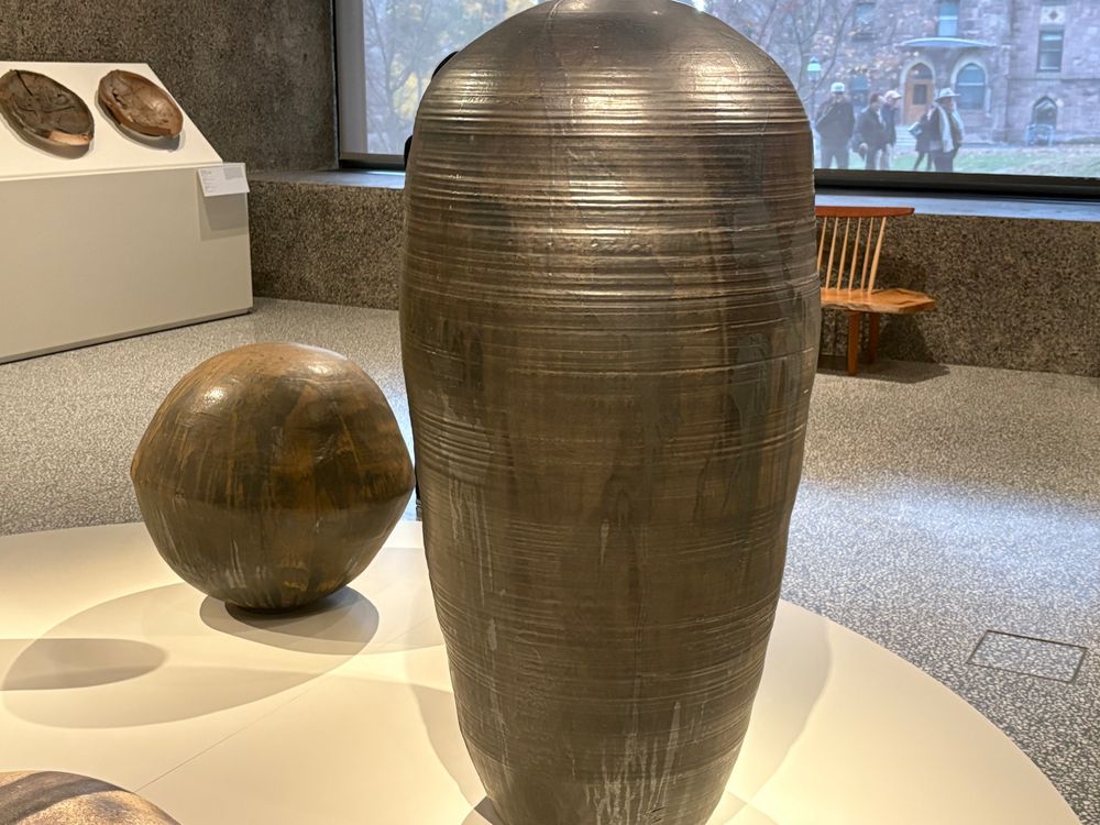 Large urn like pot with fine horizontal grooves, metallic bronze color.  Smaller, walnut shaped sculpture on left.