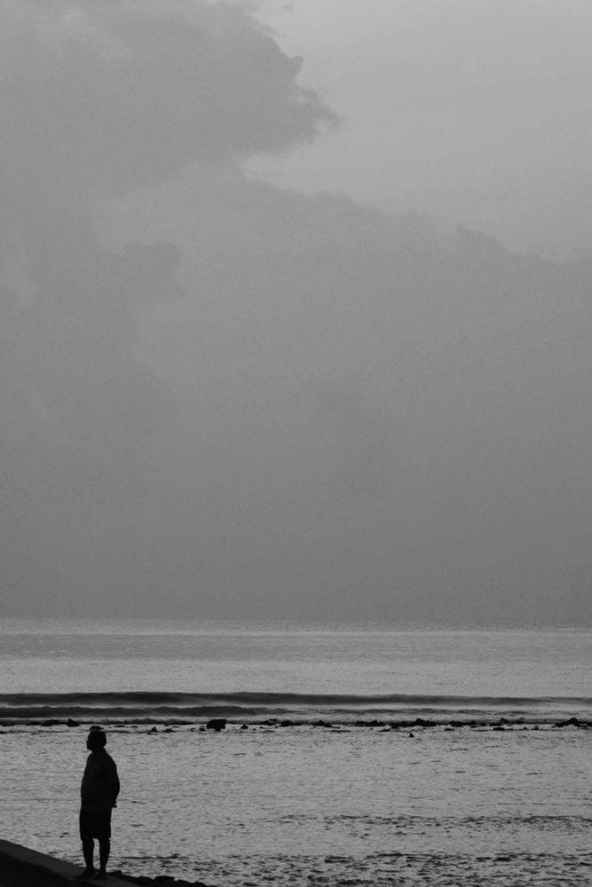 Hulhumalé, Maldives. Phase 2 sea wall. A man in silhouette is standing on the edge and instead of looking out to the sea he’s turned towards the shore looking inland. Behind him is the lagoon, reef and the sea. The sky is covered by clouds apart from a small square at the top right of the frame. The photo is in black and white. 