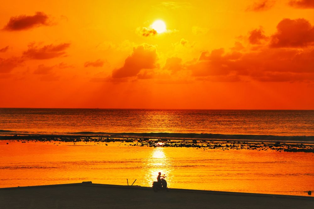 Hulhumalé, Maldives. Sunrise Golden Hour on the Hulhumalé 2nd Phase Bridge. Looking out at the lagoon and the sea. At the foreground is the breakwater with a single motorcyclist sitting at the edge watching the rising sun, centered in the brightest ray of the sun. Then the lagoon which is glass like calm with the golden rays reflecting off it. Beyond it is the reef with the corals showing at low tide and then a bit rougher sea with a more darker reddish color. The sky is dreamed by a man upside down semi circle of clouds lit up in light tangerine. Framed in the middle open part of the semi circular cloud formation is a golden sun lighting the clear sky surrounding it with gold.