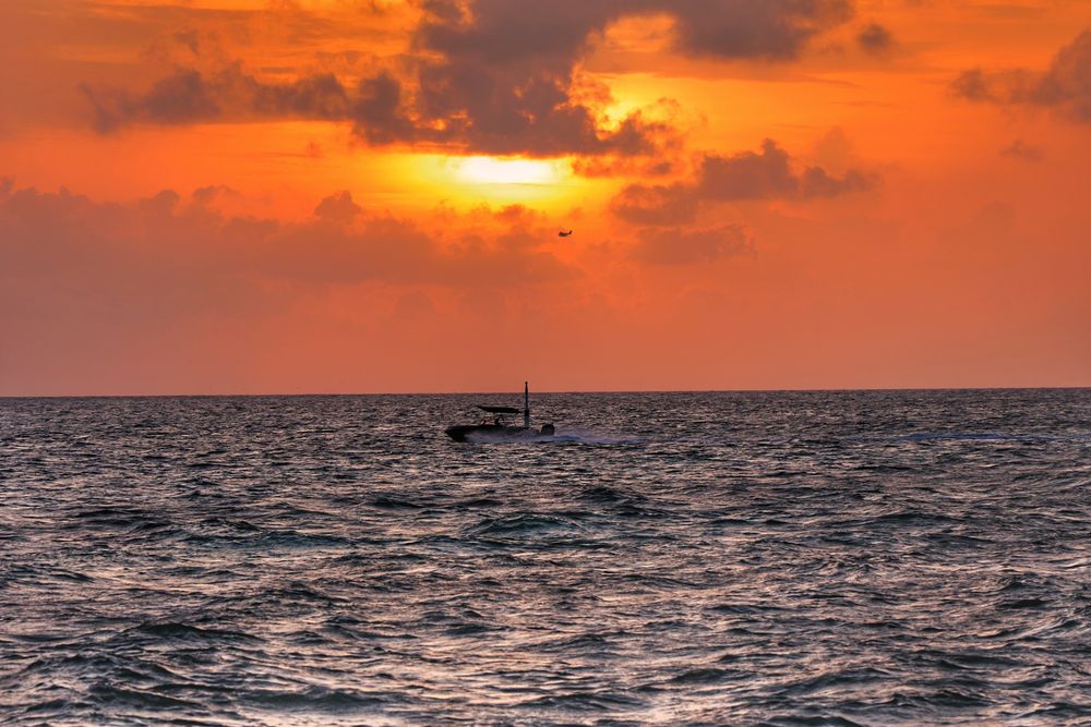 Thulusdhoo, Maldives. Sunset Golden Hour at the Naafi Jetty. In the foreground, the lagoon exit is visible, with choppy, dark blue waters. A small speedboat is positioned at the same level as a reef marker, hurrying into port before the sun sets. The sky above is painted in turquoise hues, with a golden glow surrounding the setting sun in the center. Dark gray clouds frame the sun and its radiant halo. In the distance, just below the sun, a small seaplane can be seen flying off into the sunset. 