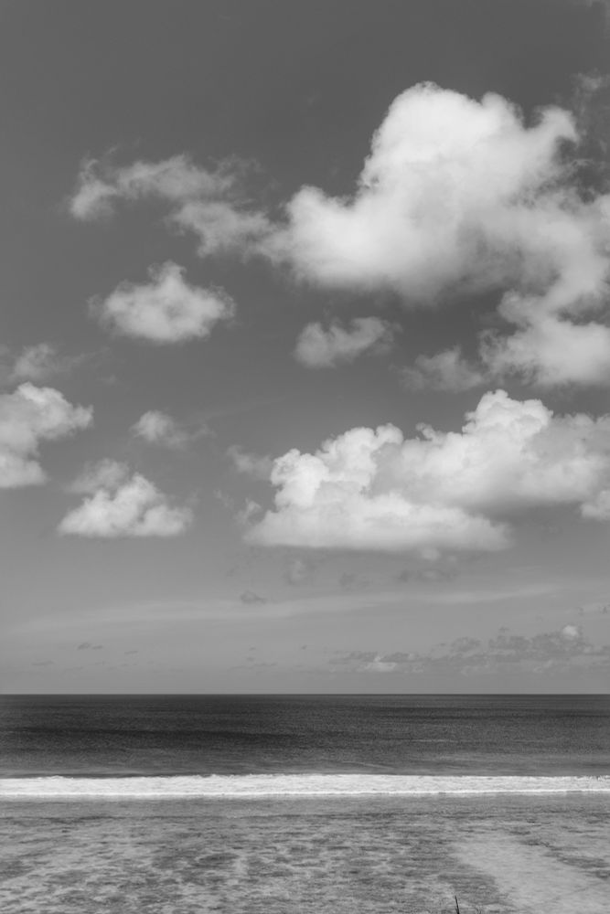 Hulhumalé, Maldives. A black and white photo of Hulhumalé beach from the balcony. The foreground is the light color of the lagoon, then the white of the waves breaking on the reef to the darker sea. The sky is scattered with puffy white clouds. The photo was taken just after noon. 