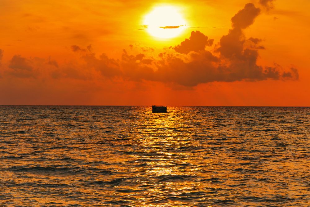 Thulusdhoo, Maldives. Sunset Golden Hour at Sunset Beach. The huge lagoon reflects the golden rays of the sun in the foreground. The water is tangerine tinted with a golden line running vertically in the middle of the scene. Mid frame is a small rectangular anchored floating object in silhouette. The sky goes from tangerine to gold. There is a horizontal cloud bank silhouetted in the glow of the sun. The golden sun is in the upper middle of the frame and is out of focus. 