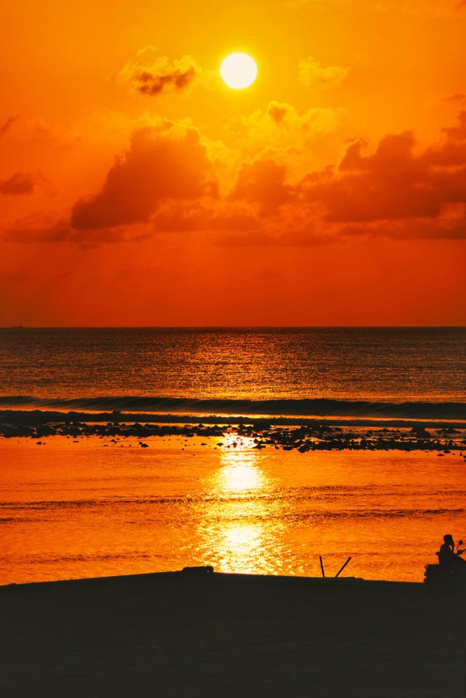 Hulhumalé, Maldives. Sunrise Golden Hour on the Hulhumalé 2nd Phase Bridge. Looking out at the lagoon and the sea. At the foreground is the breakwater with a single motorcyclist sitting at the right side edge watching the rising sun. Then the lagoon which is glass like calm with the golden rays reflecting off it. Beyond it is the reef with the corals showing at low tide and then the sea with a darker blue color. The sky is centered with a scattering of clouds backlit in tangerine. The sky below the clouds is tangerine while the sky above the sun is golden. 