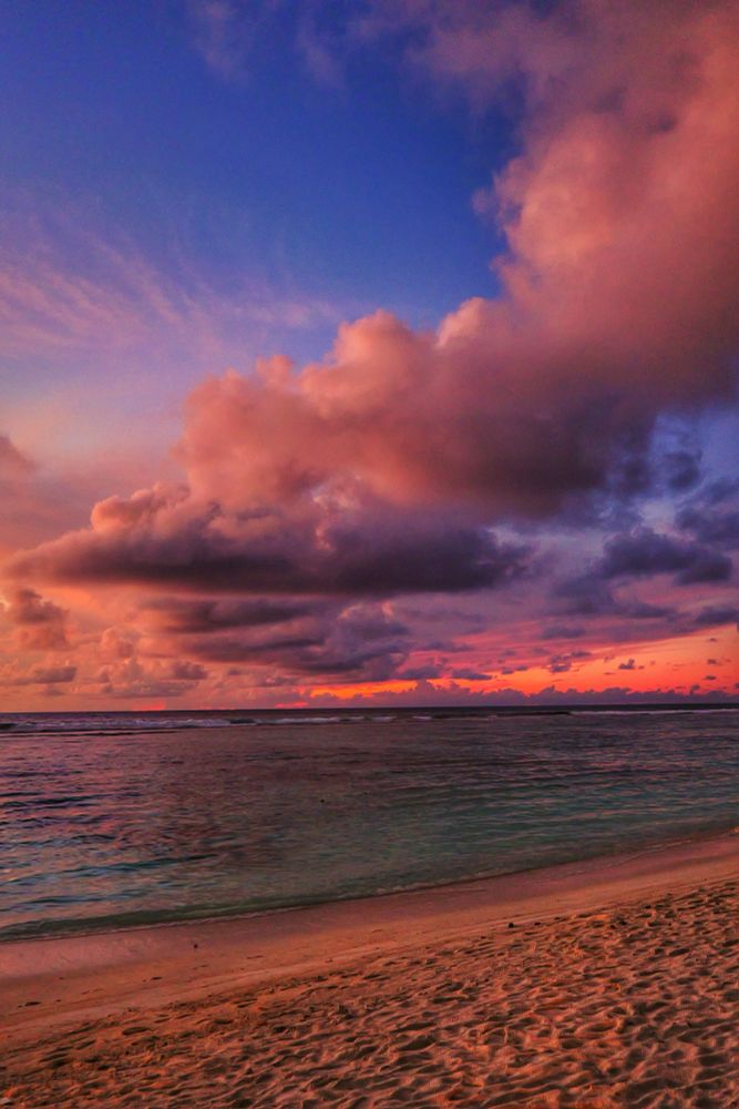Hulhumalé, Maldives. Sunrise Golden Hour at the shoreline. Sandy white beach with footsteps near the water is in the foreground. The sea is turquoise with the dark corals in the shallow showing through. The sky above the horizon is a huge snake like cloud formation coming from the background to the foreground illuminated by the rising sun. The sky below the clouds is the golden orange of the rising sun while the sky above the clouds is pure blue.