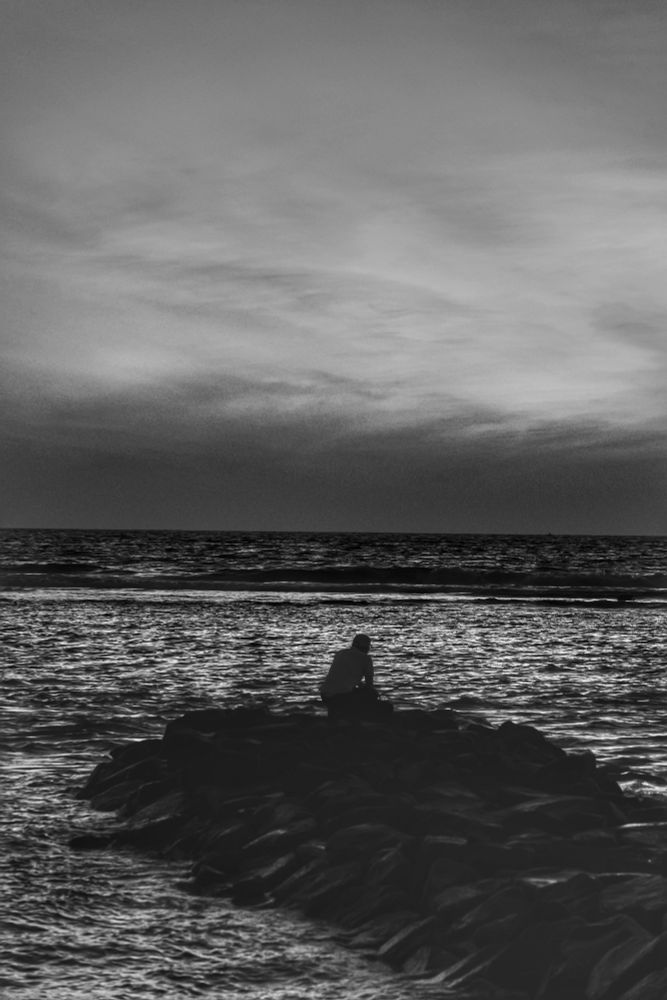 Hulhumalé, Maldives. A man is sitting down in a crouching position, looking out to the sea in contemplation on the groyne near the Hulhumalé Canal. The lagoon is lit up in silver by the sun’s rays. The sea beyond the reef is dark. There is a horizontal layer of dark clouds low near the horizon. The sky above it is mostly covered by a thin layer of light clouds. The photograph is in black and white. 