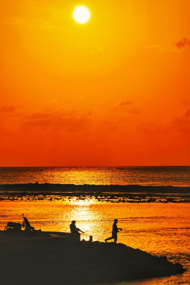 Hulhumalé, Maldives. Sunrise at the 2nd Phase sea wall. In the foreground is a parked scooter. Beyond that there are two people, one sitting on the sea wall boundary and another standing on the rocks and using a rod to fish in the lagoon. Everything is in silhouette. The lagoon is in gold. The reef is in low tide and the corals are showing above water and beyond that the waves crash. Both there too are in silhouette. The sea is simmering in gold and black. The clods in the sky are very faint and some nearly translucent. The sun is shining top center left of the frame. The color around the sun is gold and it darkens to tangerine as it goes further from the sun in a circular fashion. Light halos can be seen around the sun. 