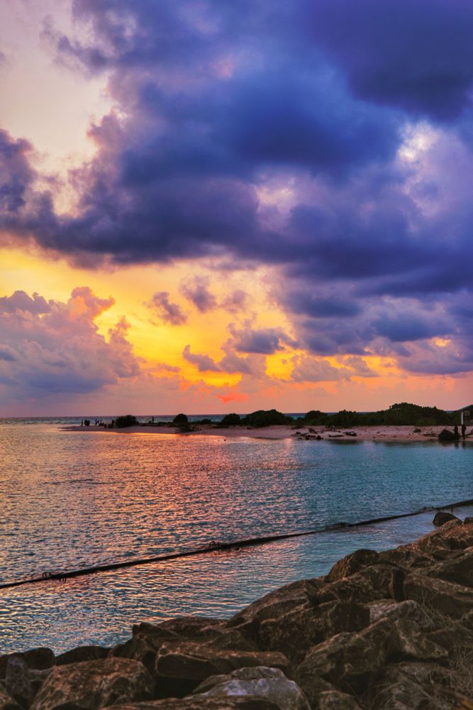 Thulusdhoo, Maldives. Sunset Golden Hour at the beach looking at the Thulusdhoo Sunset Beach in the distance protruding out into the lagoon. At the foreground is the rocks put to stop erosion at certain parts of the island’s beach. Then the turquoise lagoon cut in half by a beam of golden reflection from the sun’s rays. Beyond that is the strip of land jutting out of the island called the Sunset Beach and the best spot to view one. It has no large trees, just shrubs. Beyond that the horizon is lit in soft orange to light gold near the low sun. Above it the light is cut by clouds. One huge dark cloud comes in from the upper right of the frame and dominates the whole blue sky. 
